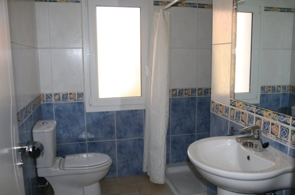 Cuarto de baño con azulejos en tonos azul y blanco, equipado con lavabo de pedestal, inodoro y plato de ducha con cortina e iluminación natural.