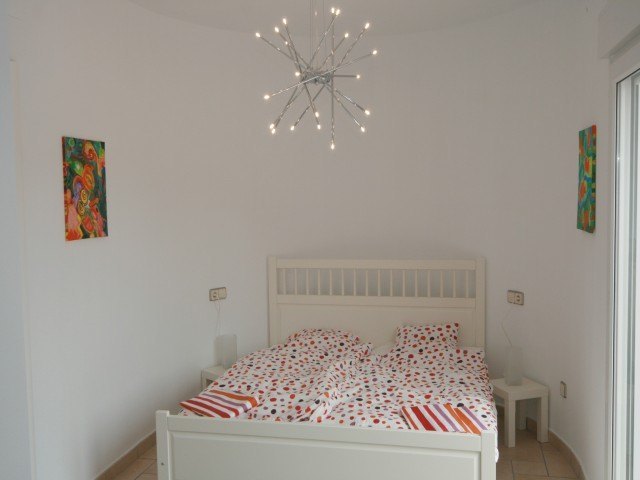 Dormitorio con cama de madera blanca, lámpara de techo de diseño contemporáneo y paredes blancas decoradas con arte colorido.