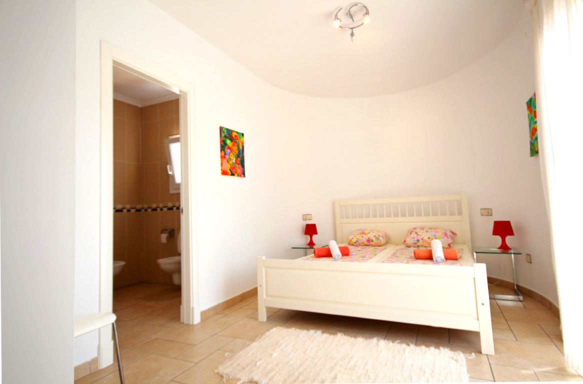 Dormitorio con paredes curvas, cama de madera blanca y acceso directo a un baño privado con azulejos en tonos tierra.