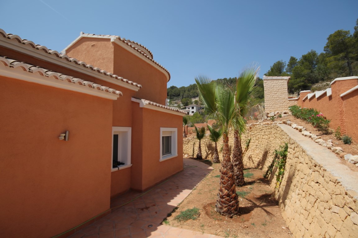 Fachada de color terracota con carpintería blanca, flanqueada por un camino pavimentado y muros de piedra seca con palmeras jóvenes.