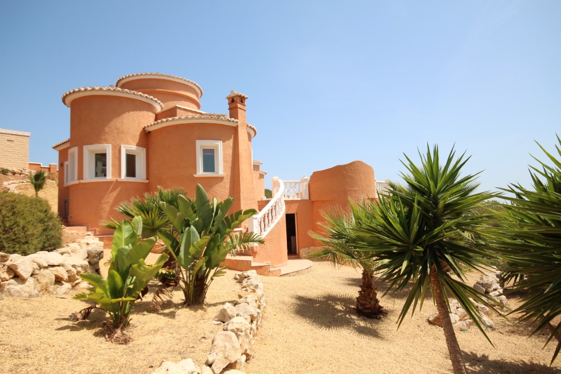Fachada de formas circulares en tono terracota con escalera exterior de balaustrada blanca y jardín mediterráneo de grava con yucas y palmeras.