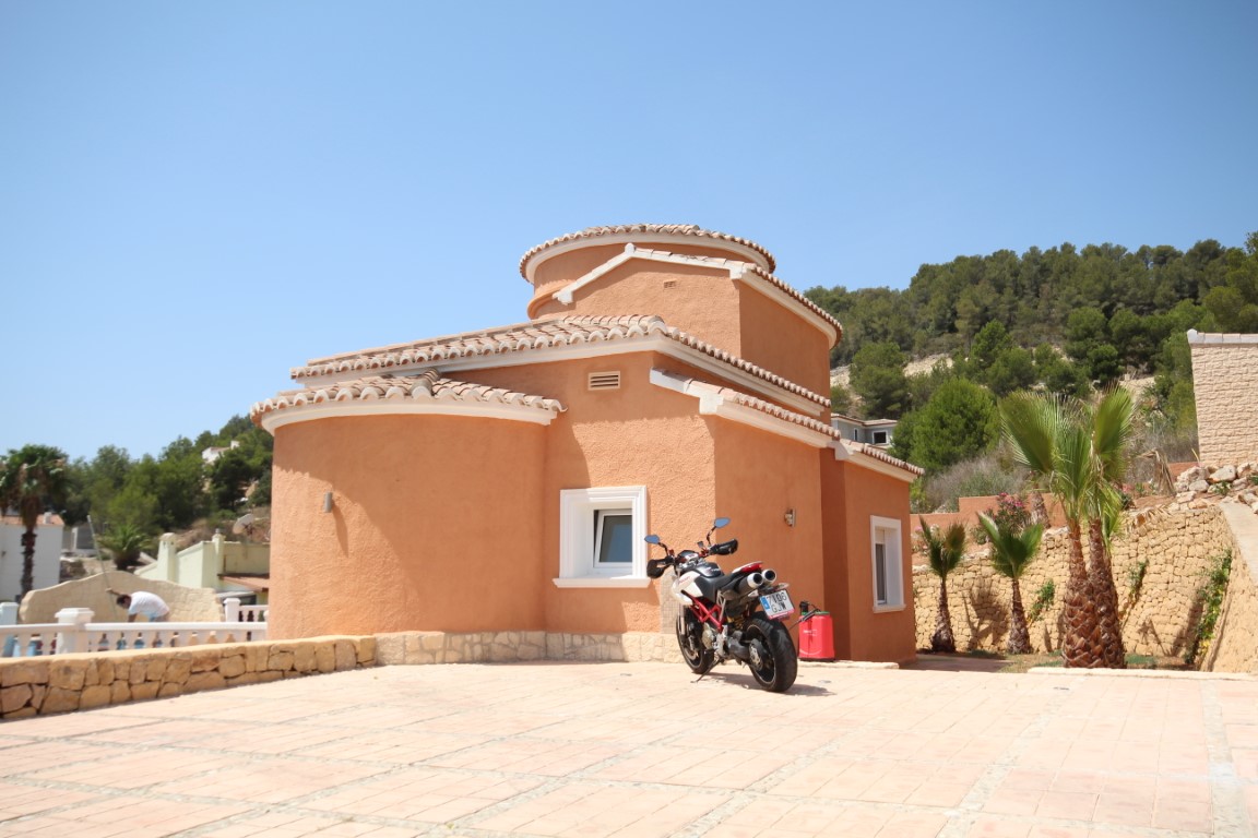 Fachada mediterránea con muros curvos en tonos tierra, amplia zona de aparcamiento pavimentada y muros de piedra natural con palmeras.