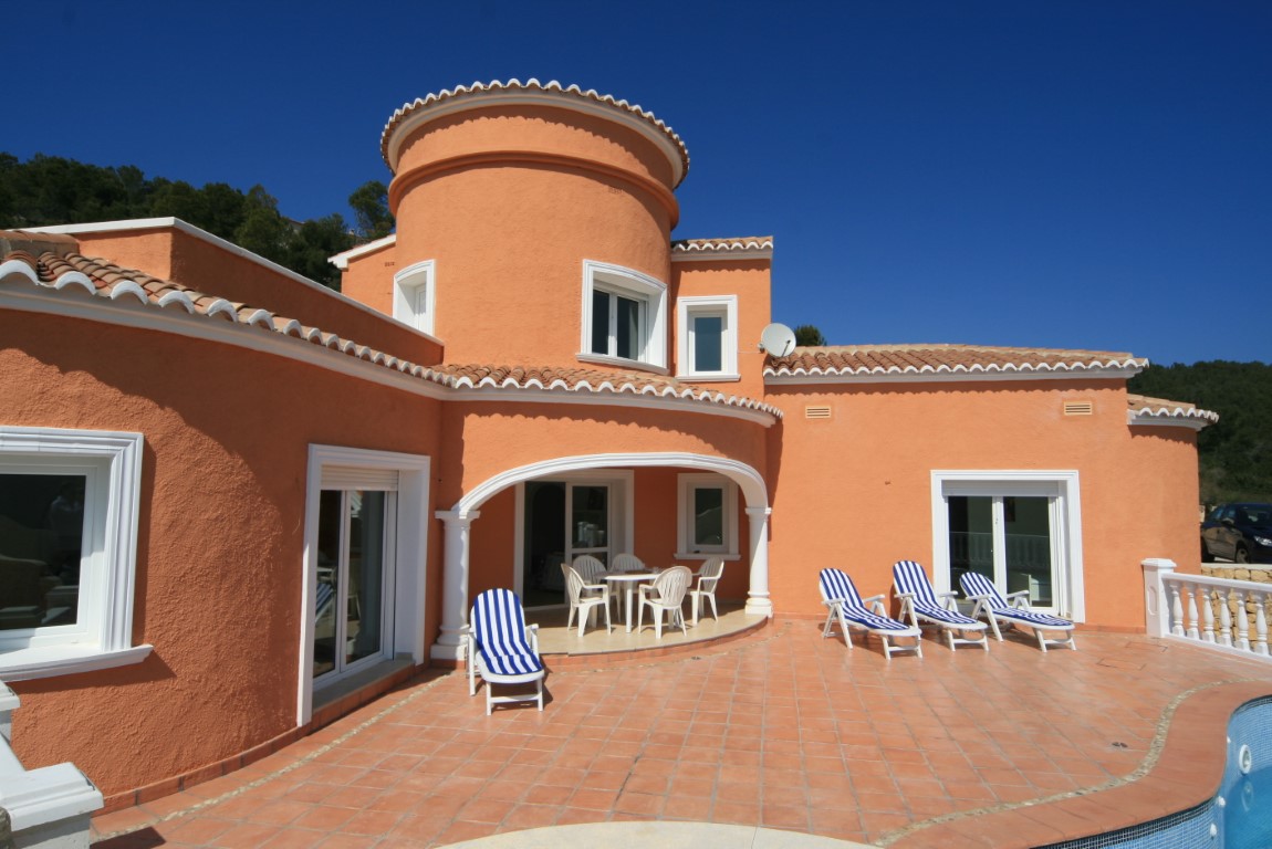 Fachada mediterránea en tonos terracota con torre circular, porche arqueado y amplia terraza de gres junto a la piscina con tumbonas.
