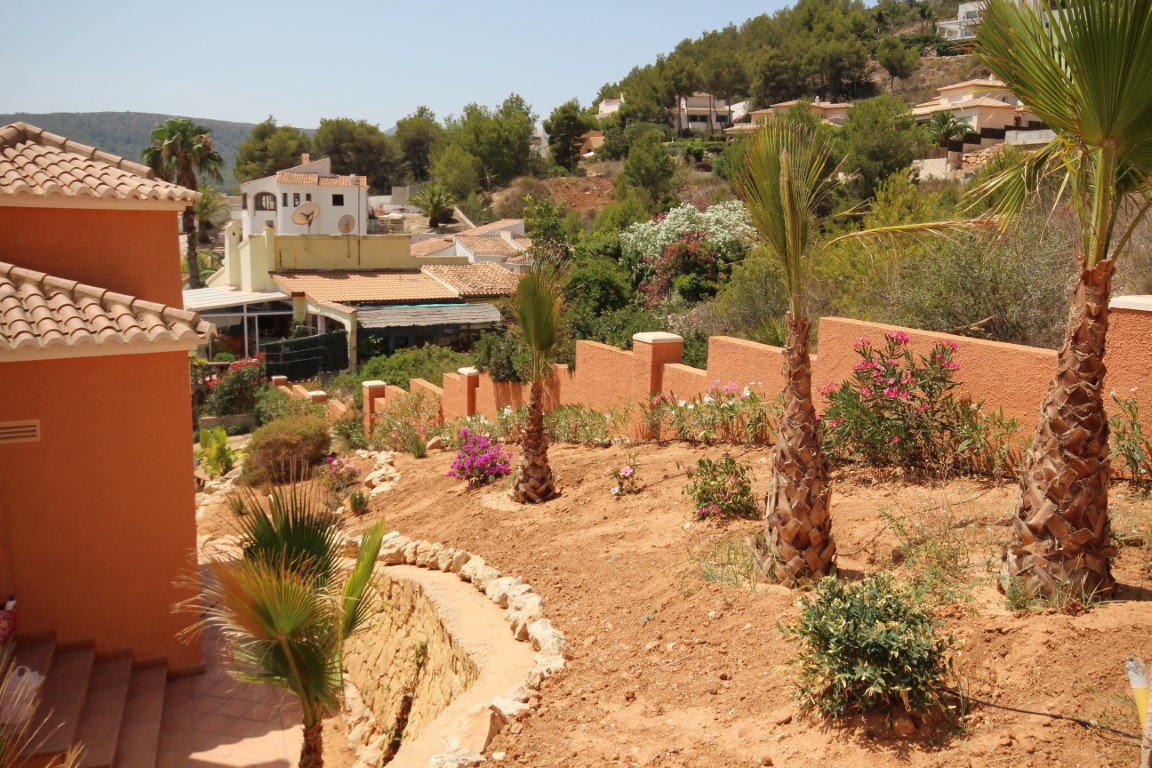 Jardín mediterráneo aterrazado con palmeras jóvenes, adelfas y muros perimetrales en tonos terracota, integrado en una urbanización con vistas despejadas.