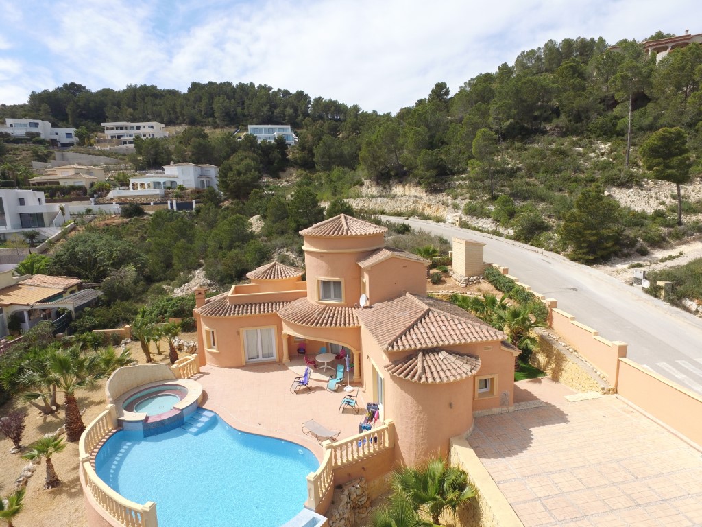 Vista aérea de la villa con fachada terracota, destacando la piscina de forma orgánica con jacuzzi integrado y la amplia terraza solárium rodeada de vegetación mediterránea.