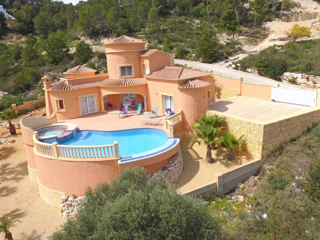 Vista exterior de la villa con piscina de borde infinito, jacuzzi integrado y amplia terraza. Incluye zona de aparcamiento privada y muros de piedra natural.