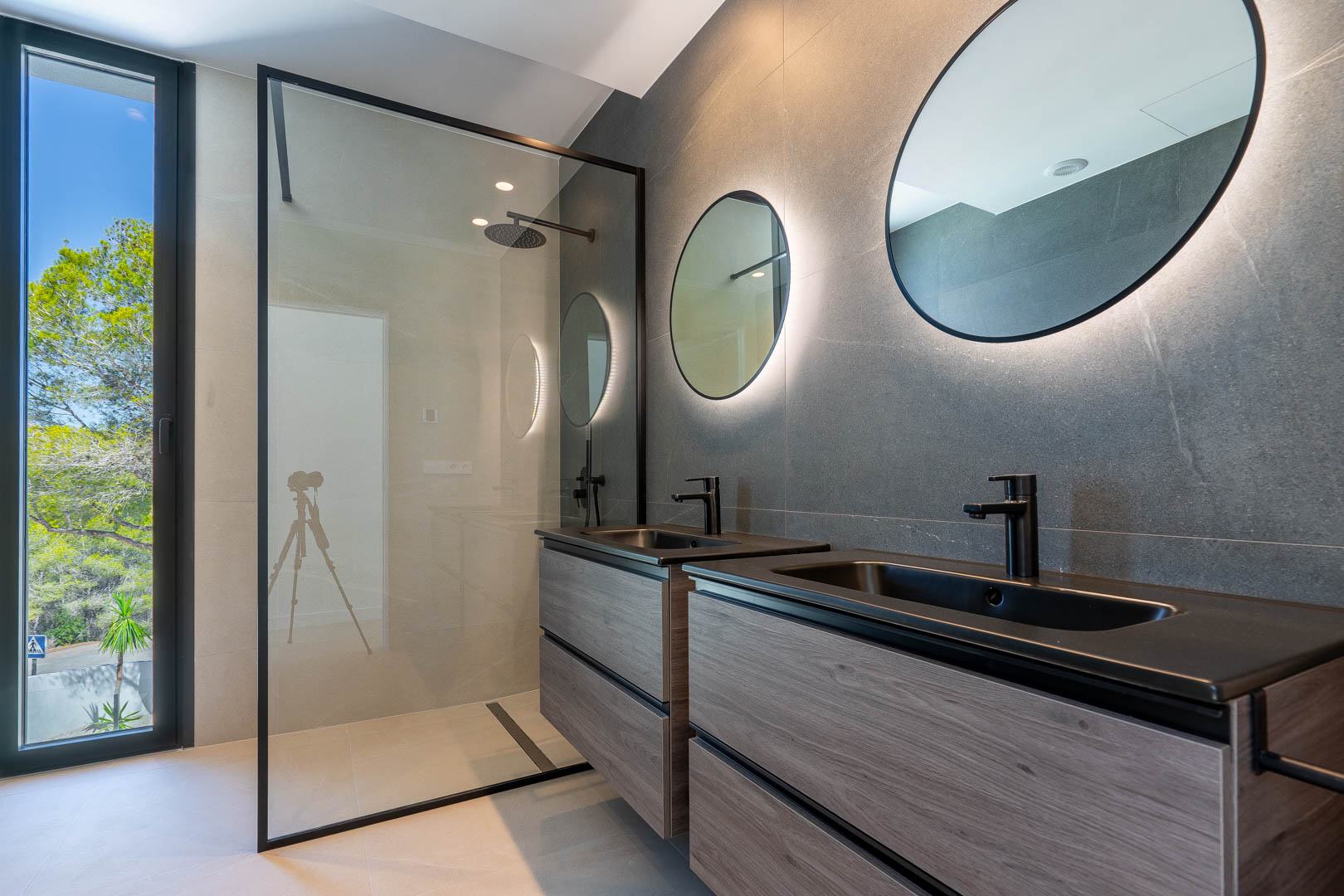 Baño de diseño con lavabo doble en madera, espejos circulares retroiluminados y ducha italiana con mampara de cristal y grifería negra mate.