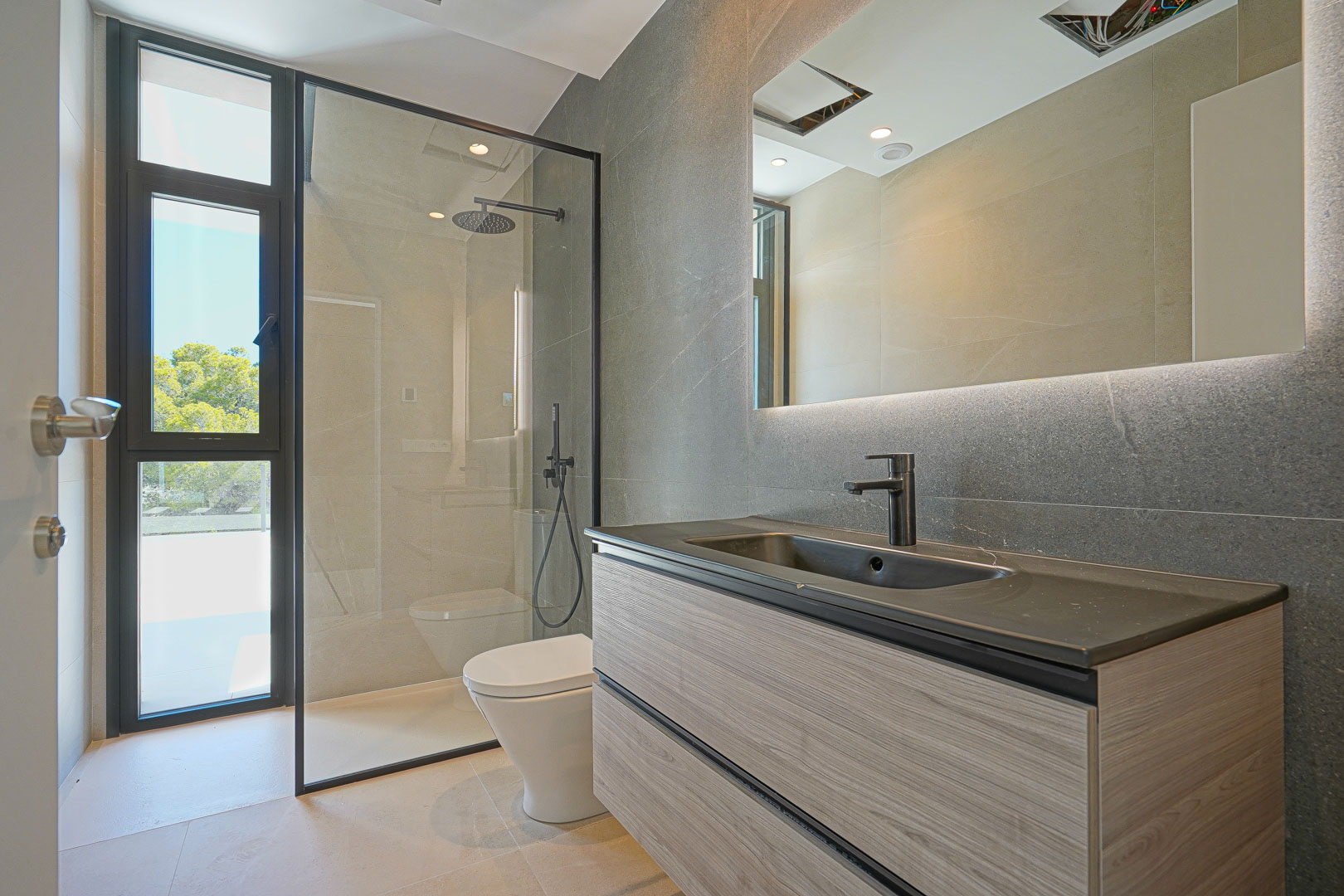 Baño moderno con mueble suspendido de madera, lavabo oscuro, espejo retroiluminado y ducha italiana con mampara de perfil negro y vistas al exterior.