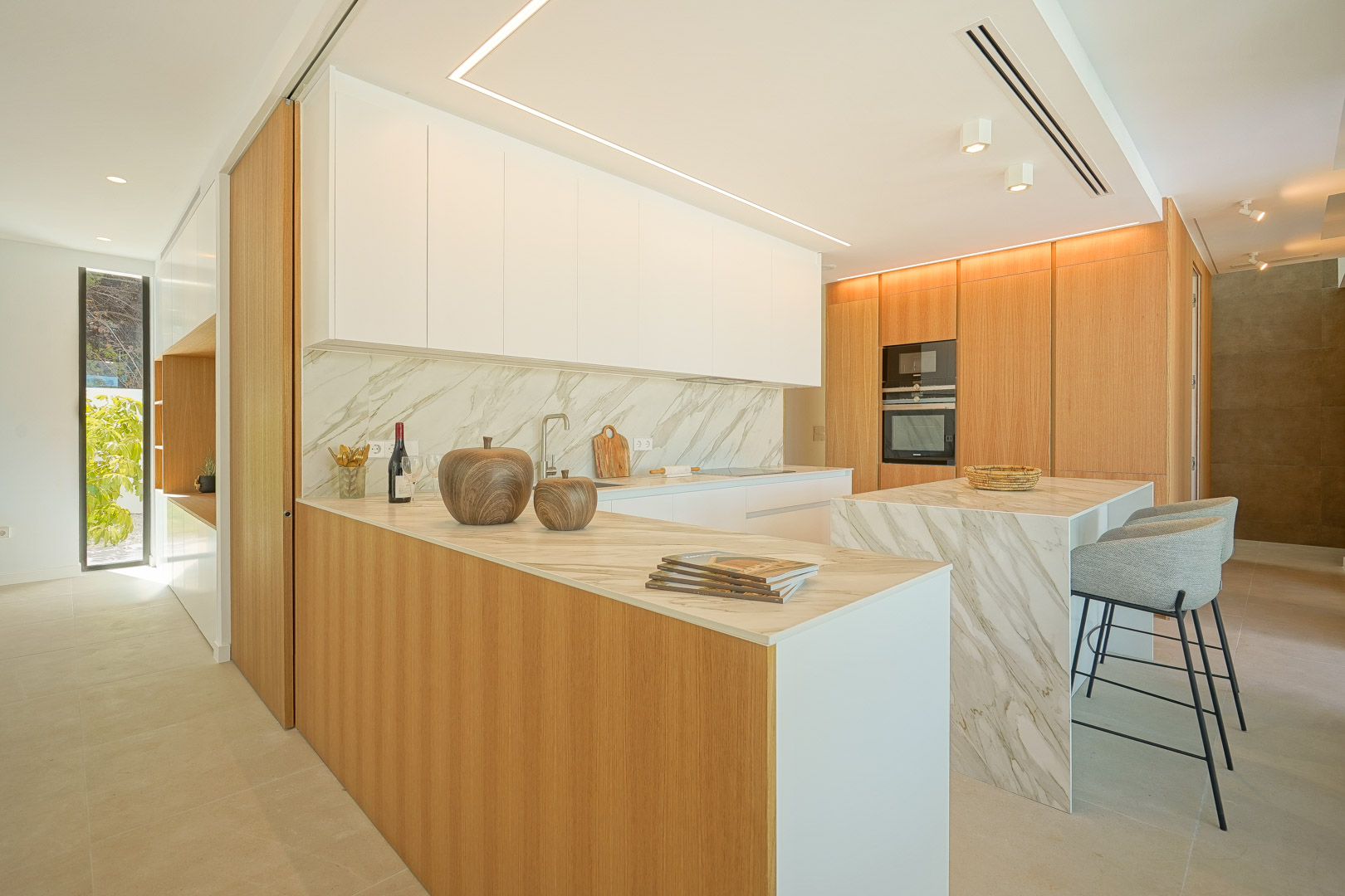 Cocina americana con encimeras de mármol, armarios de madera de roble y una isla central con taburetes. Incluye electrodomésticos integrados e iluminación LED lineal en el techo.