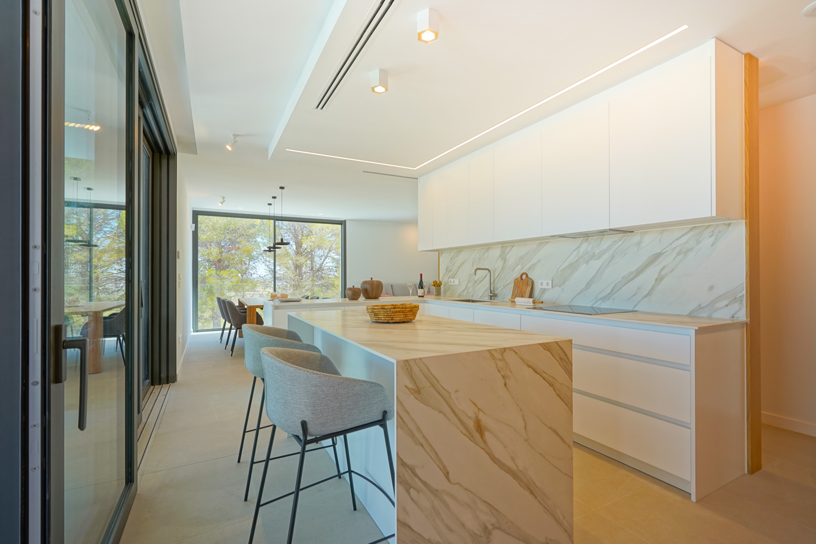 Cocina americana con isla de efecto mármol, taburetes grises y armarios blancos. Incluye iluminación LED integrada y acceso directo a la terraza exterior.