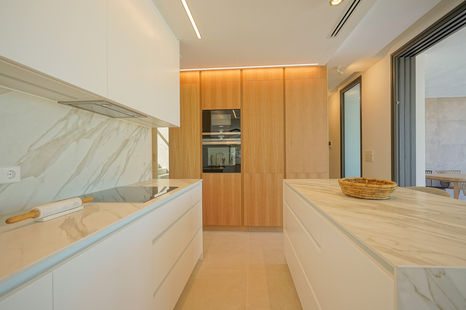 Cocina moderna con encimeras de mármol veteado, armarios blancos minimalistas y una pared de almacenamiento en madera con electrodomésticos integrados e iluminación lineal.