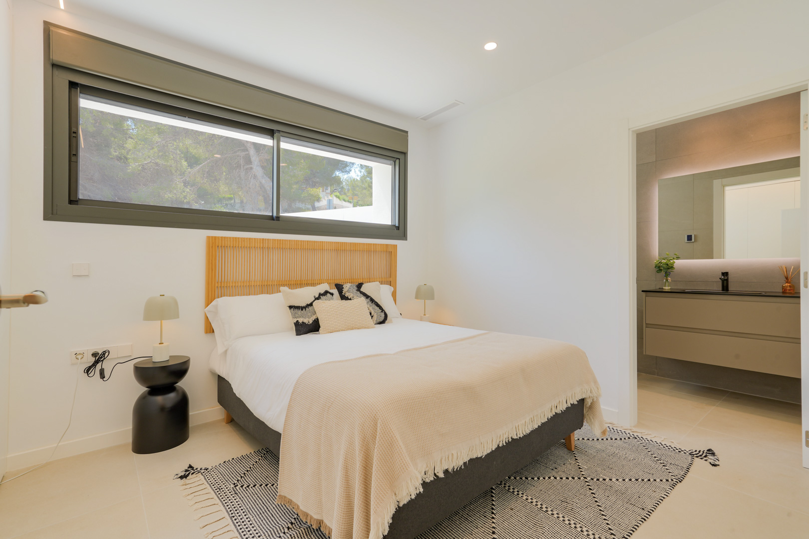 Dormitorio doble con cabecero de madera, ventana panorámica con vistas al entorno boscoso y acceso directo a un baño en suite con lavabo moderno y espejo retroiluminado.
