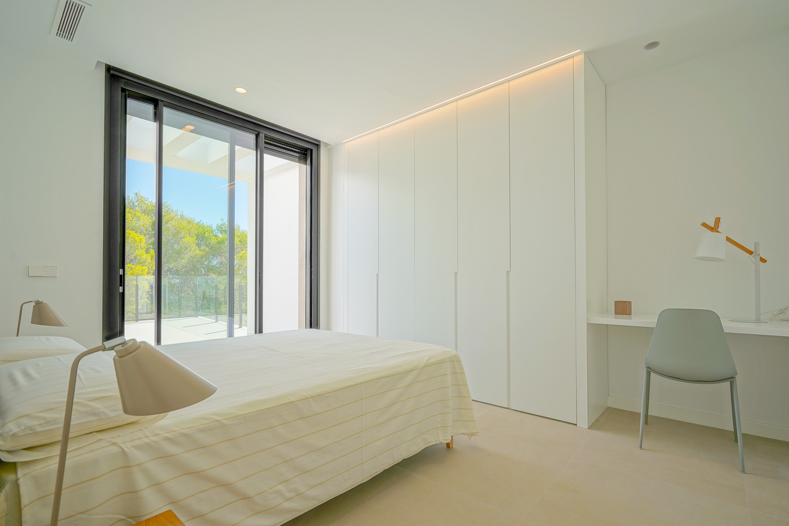 Dormitorio luminoso con armarios empotrados blancos, iluminación LED integrada, zona de escritorio y acceso directo a la terraza con vistas al bosque.