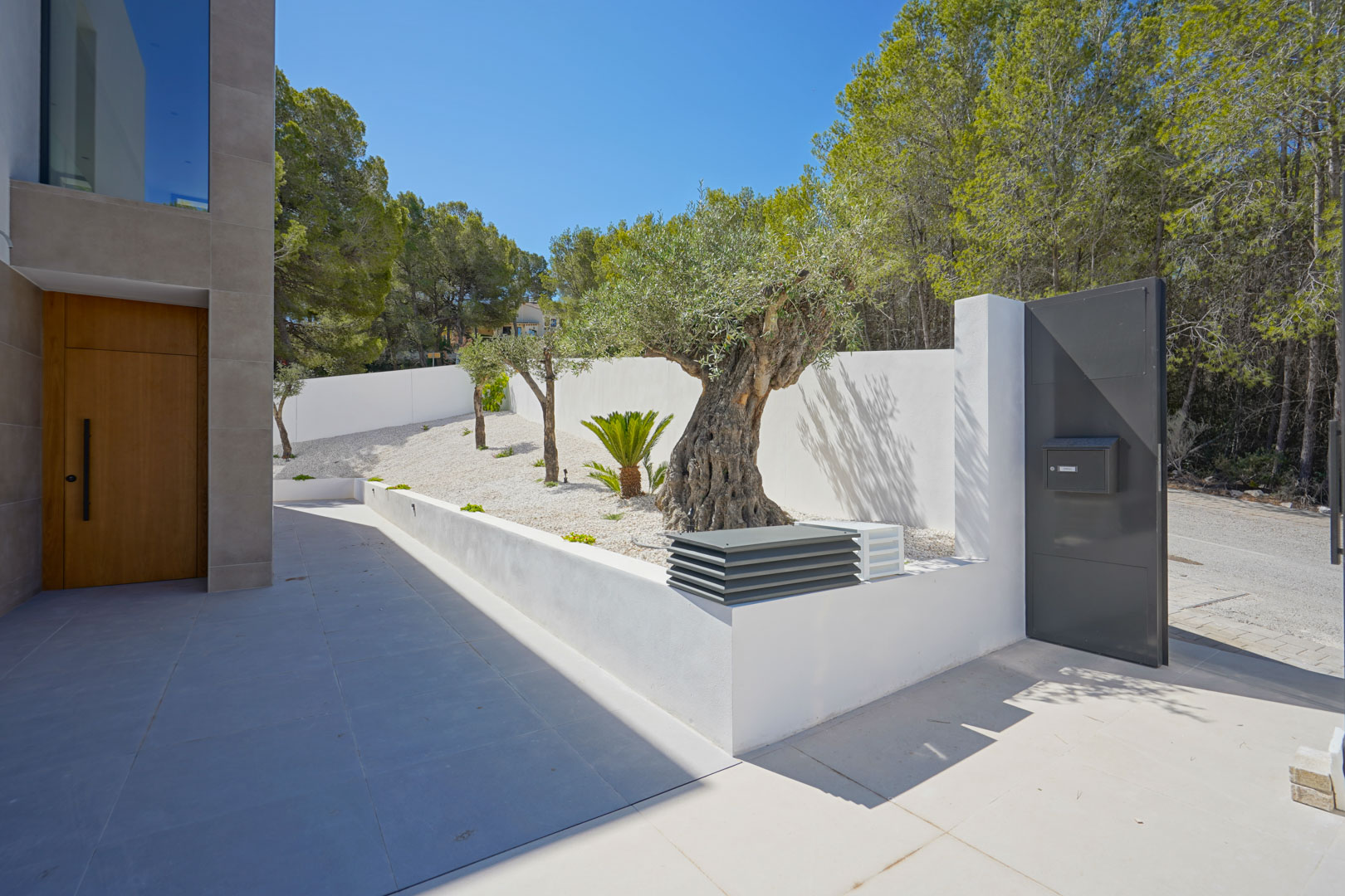Entrada principal con puerta de madera, jardín de grava blanca con olivos centenarios y muro perimetral blanco junto al acceso peatonal metálico.