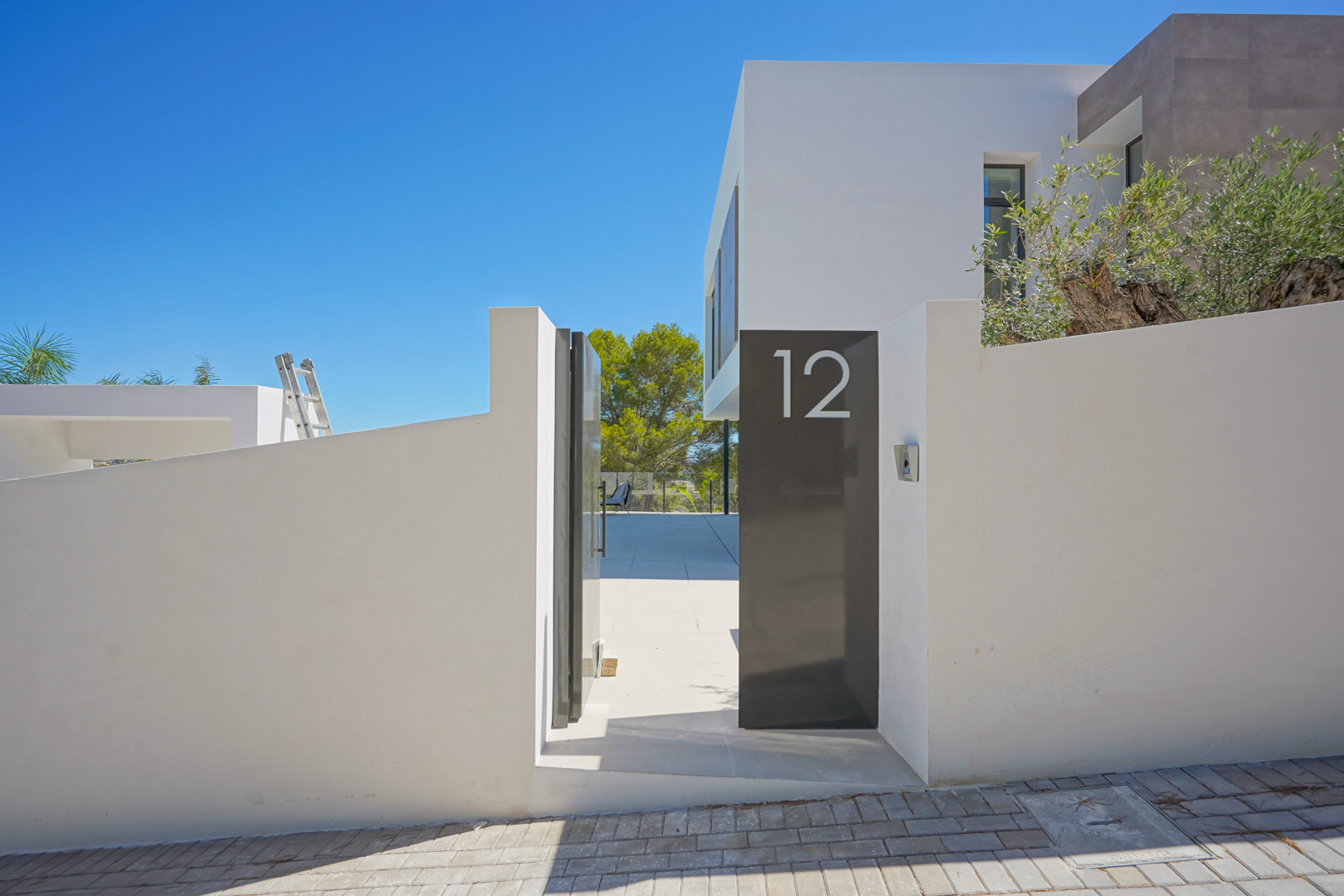 Entrada principal moderna con muro perimetral blanco, puerta peatonal metálica en gris oscuro con el número 12 y acceso a un patio pavimentado bajo el cielo despejado.