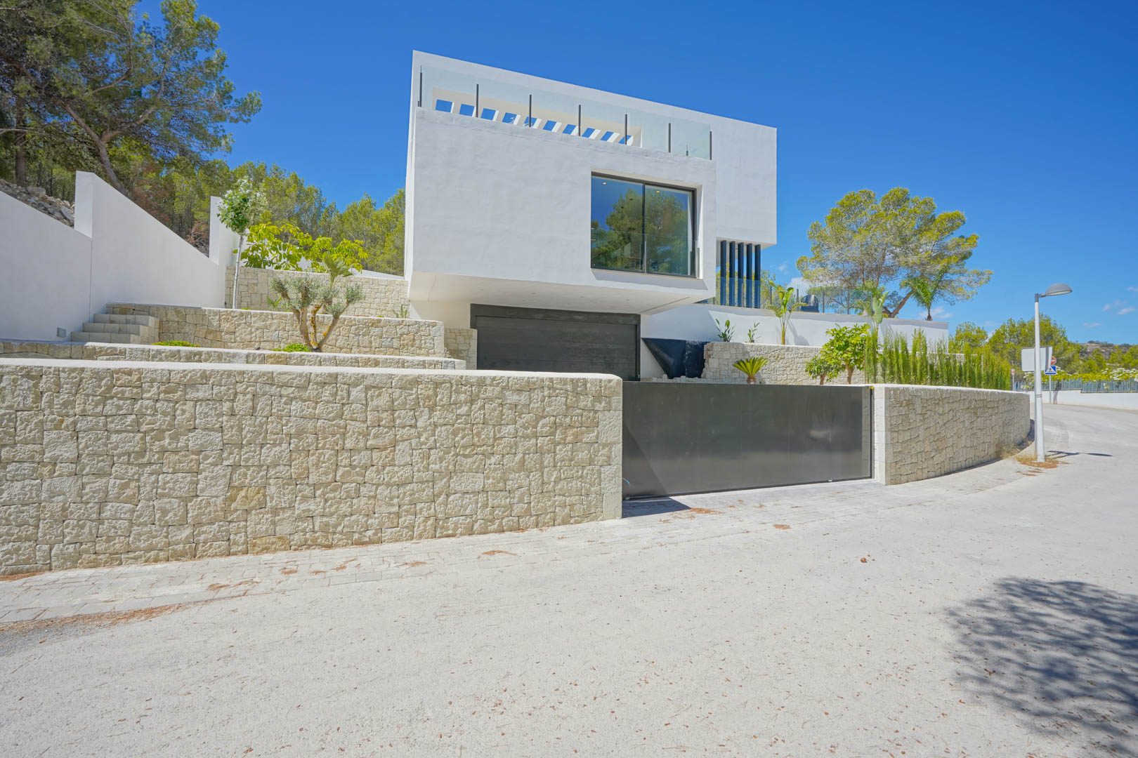Fachada exterior moderna con muros de piedra natural, acceso al garaje doble y jardín aterrazado bajo un cielo despejado en Moraira.