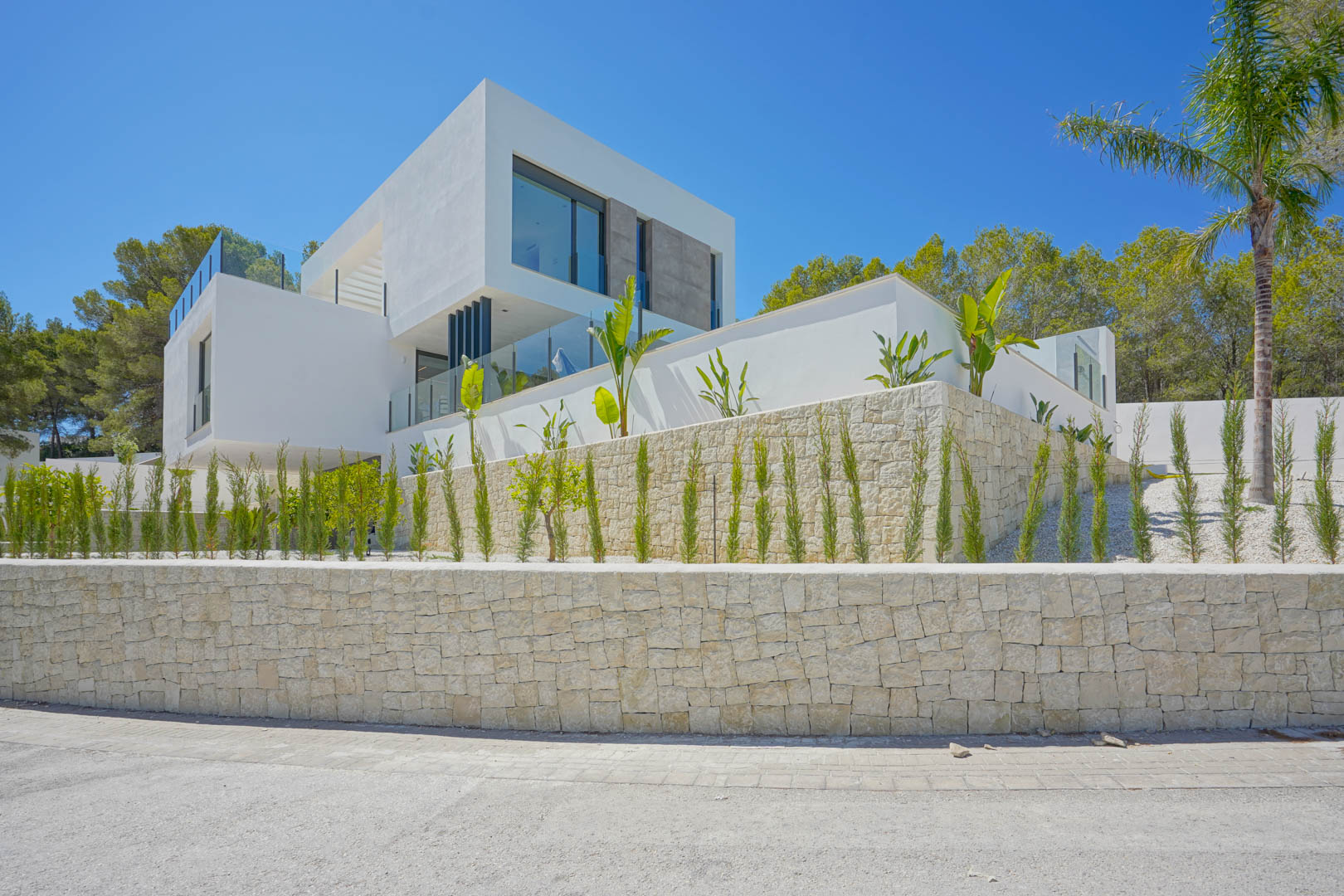 Fachada moderna de líneas rectas con muros de piedra natural, amplios ventanales y jardín mediterráneo perimetral con cipreses y palmeras bajo un cielo despejado.