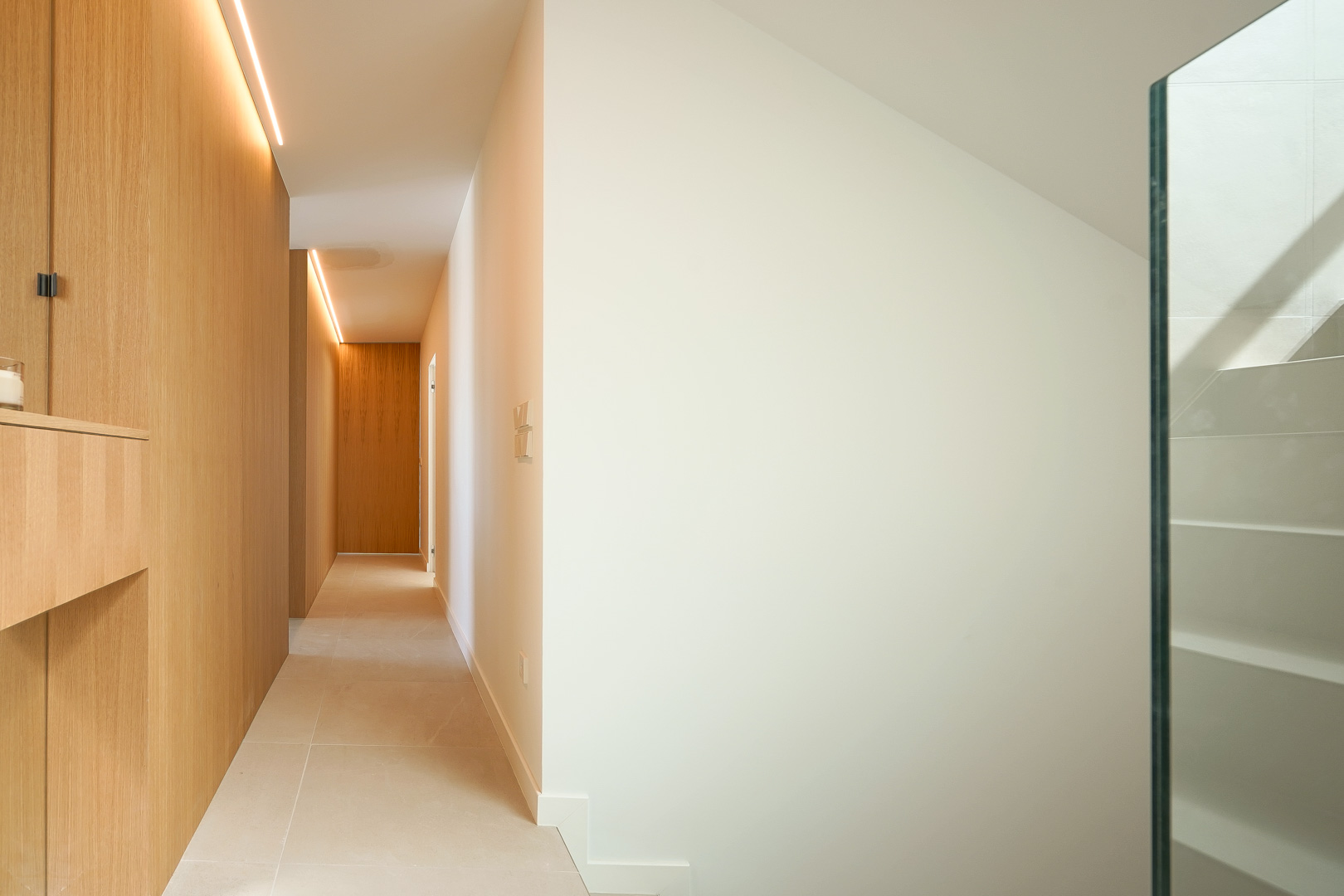 Pasillo minimalista con paneles de madera de roble e iluminación LED integrada, suelo cerámico claro y escalera con barandilla de cristal.