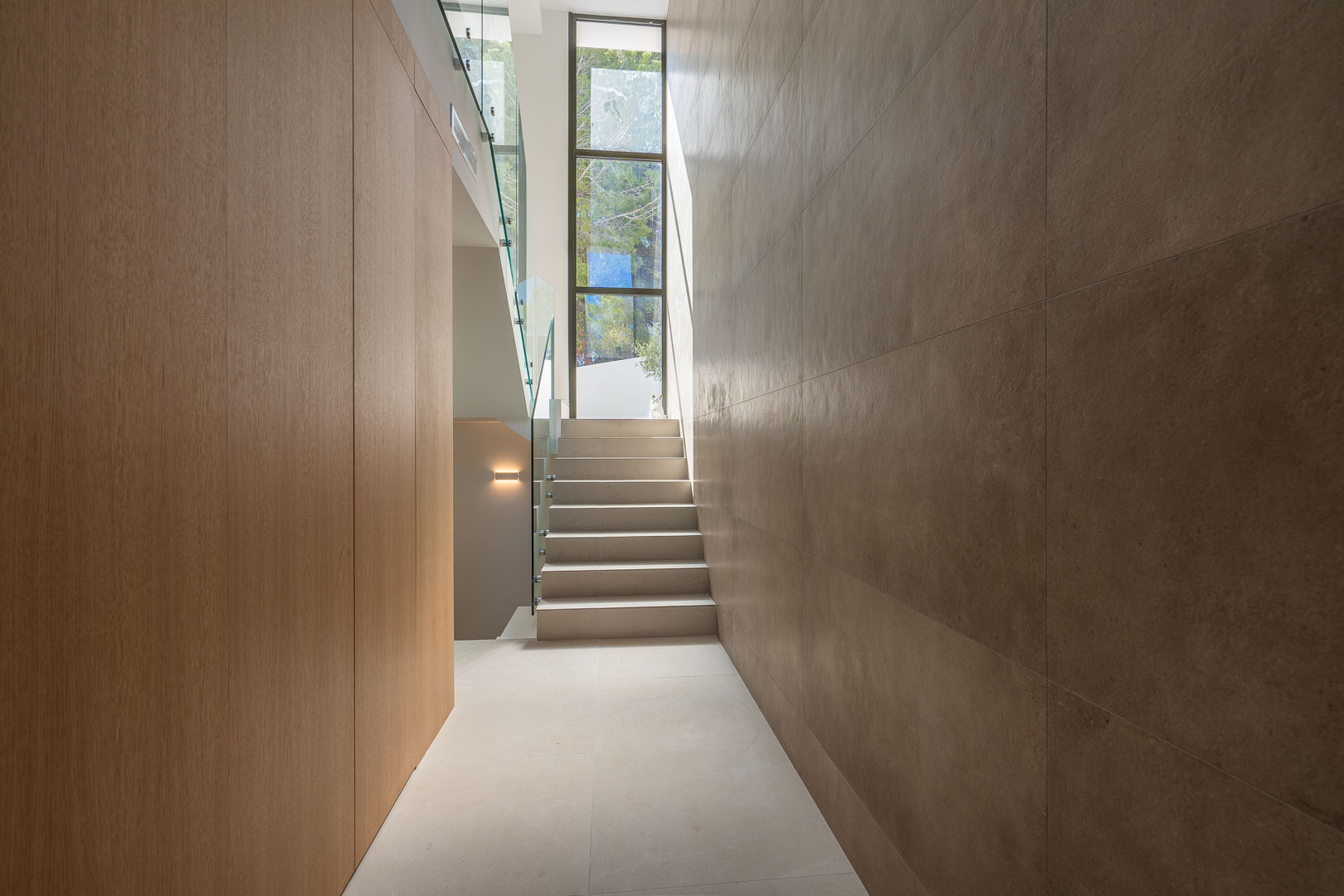 Pasillo moderno con paneles de madera, paredes de piedra de gran formato y escalera con barandilla de cristal bajo un ventanal vertical.