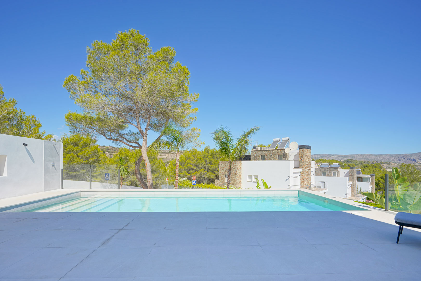 Piscina infinita con revestimiento blanco y terraza de gres porcelánico de gran formato, rodeada de pinos y vistas despejadas al entorno natural de Moraira.