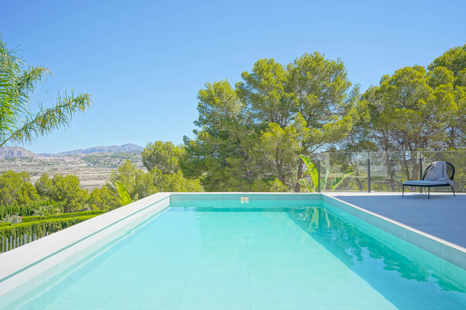Piscina rectangular de gresite claro con terraza de gres porcelánico, barandilla de cristal y vistas despejadas a la zona boscosa y las montañas de Moraira.