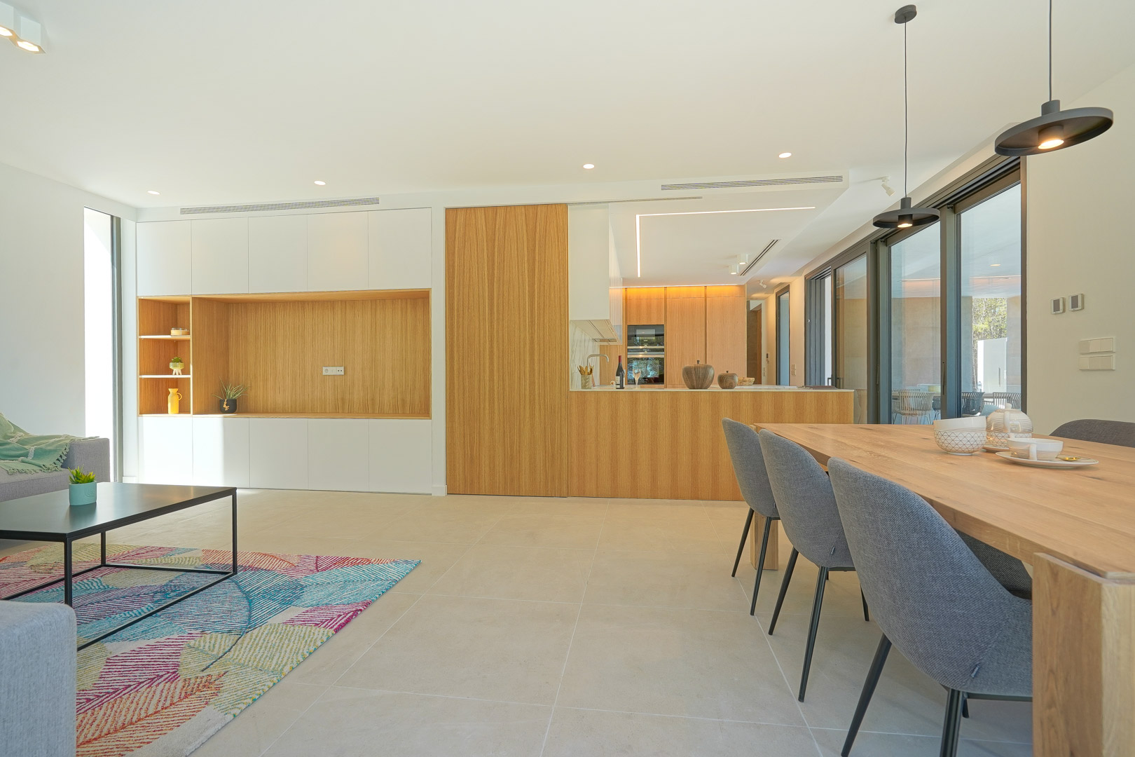 Salón-comedor diáfano con cocina integrada de madera natural, suelos de gres porcelánico y grandes ventanales que conectan con la terraza exterior.