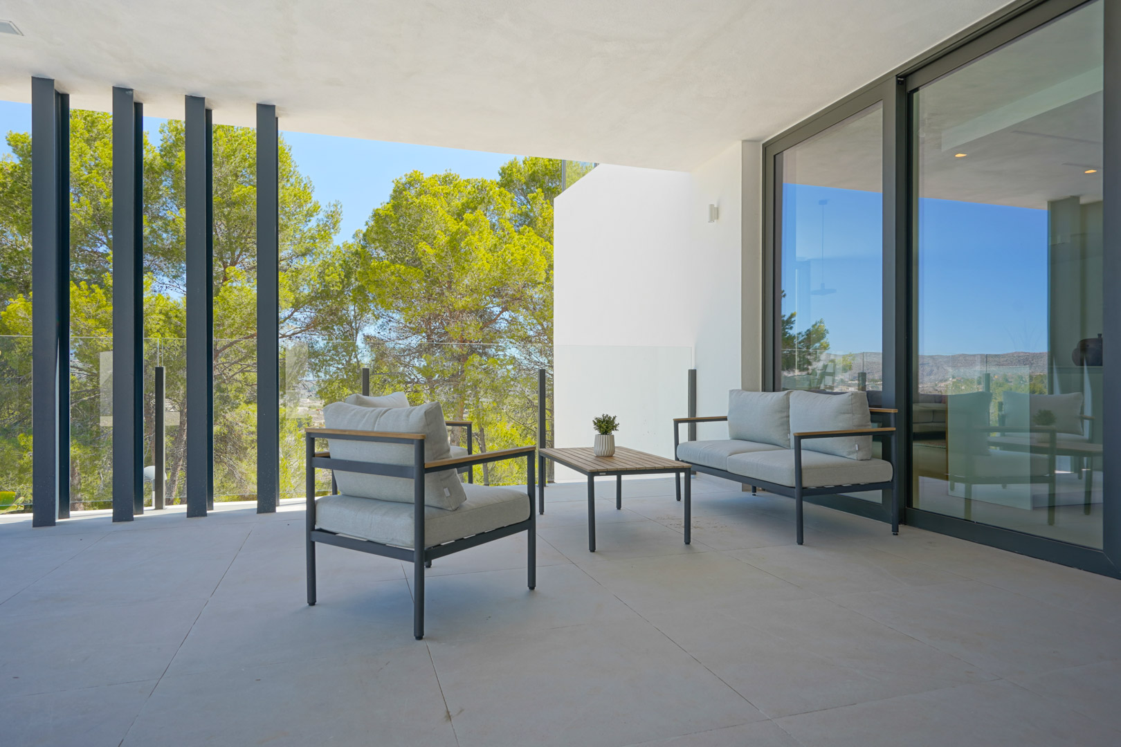 Terraza cubierta con mobiliario de exterior moderno, barandilla de cristal y vistas a la zona boscosa a través de elegantes pilares verticales.