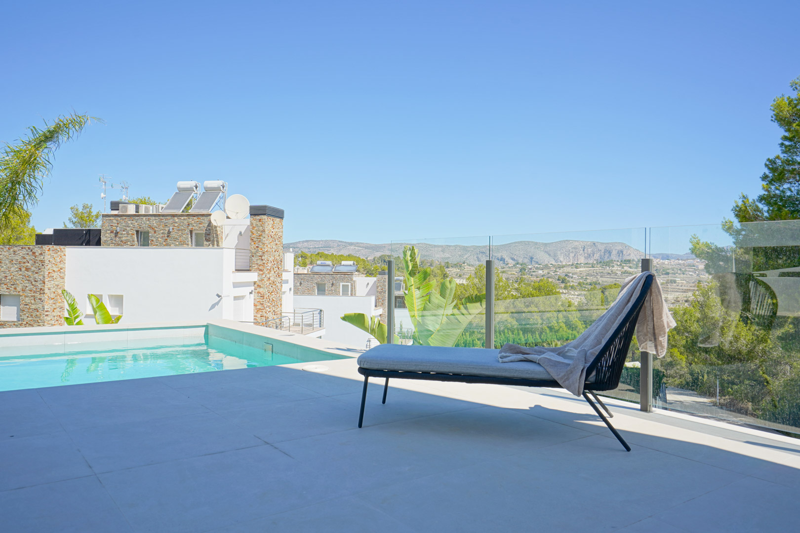 Terraza junto a la piscina con barandilla de cristal, tumbona de diseño y vistas despejadas a las montañas y el entorno boscoso de Moraira.