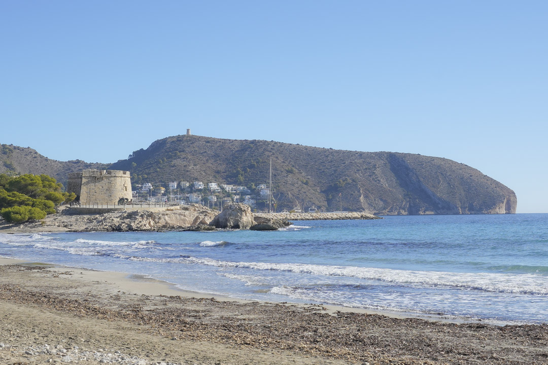 Vista de la playa de Moraira con el histórico castillo de piedra y el cabo de Cap d'Or al fondo bajo un cielo despejado.