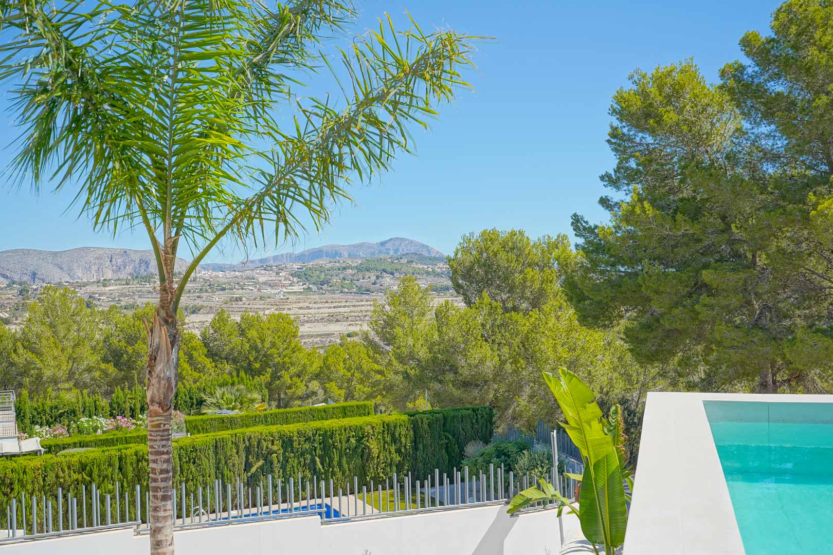 Vista panorámica desde la terraza de la piscina, destacando el entorno boscoso de pinos, setos cuidados y las montañas lejanas bajo un cielo despejado.