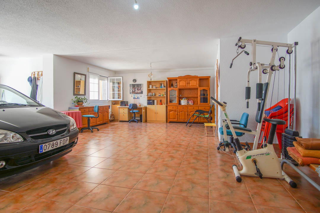 Amplio garaje en planta baja con suelos cerámicos, que integra zona de gimnasio, mobiliario de madera maciza y espacio de oficina con luz natural.