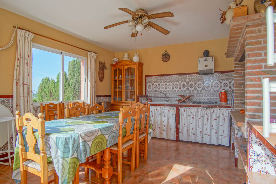 Cocina exterior cerrada con comedor, encimera de azulejos tradicionales, horno de leña de ladrillo y un gran ventanal con vistas al entorno natural.