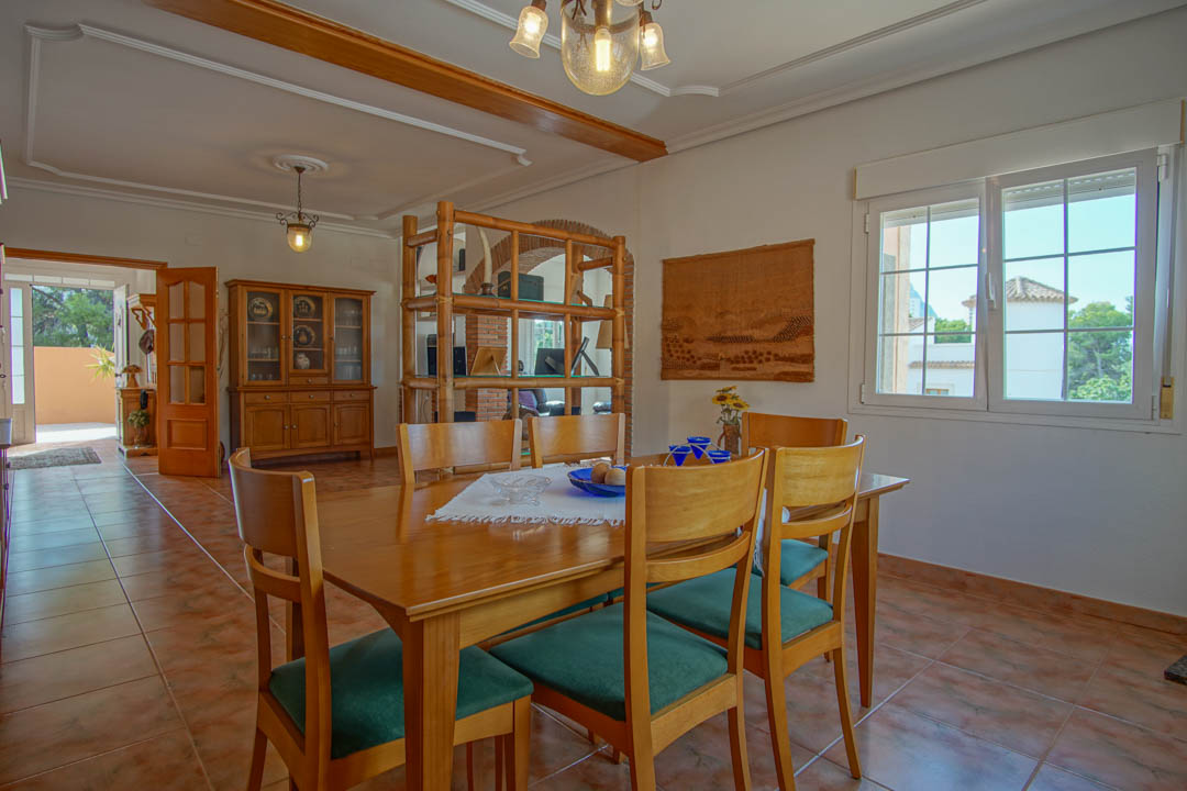 Comedor luminoso con mesa de madera maciza, sillas tapizadas en verde y suelos cerámicos. Destacan la viga de madera decorativa y la estantería que divide los ambientes.