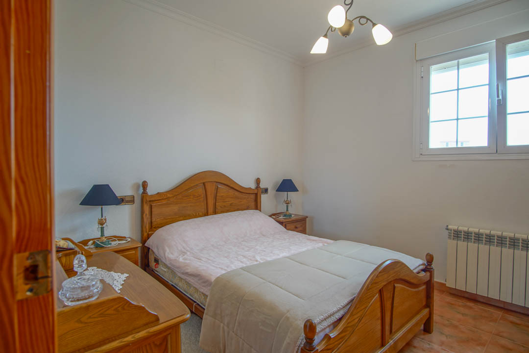 Dormitorio con mobiliario de madera maciza, suelos cerámicos y ventana de PVC. Incluye radiador de calefacción y lámparas de noche sobre mesitas de madera.