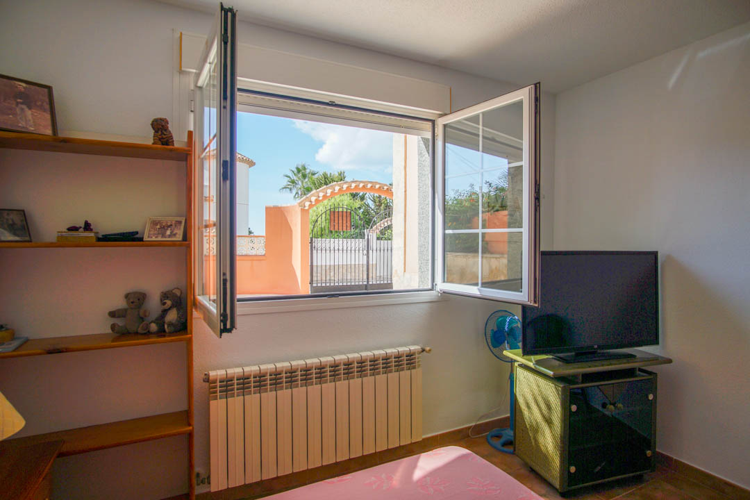 Dormitorio luminoso con estantería de madera maciza, radiador bajo la ventana y vistas al acceso exterior de la propiedad a través de carpintería blanca.