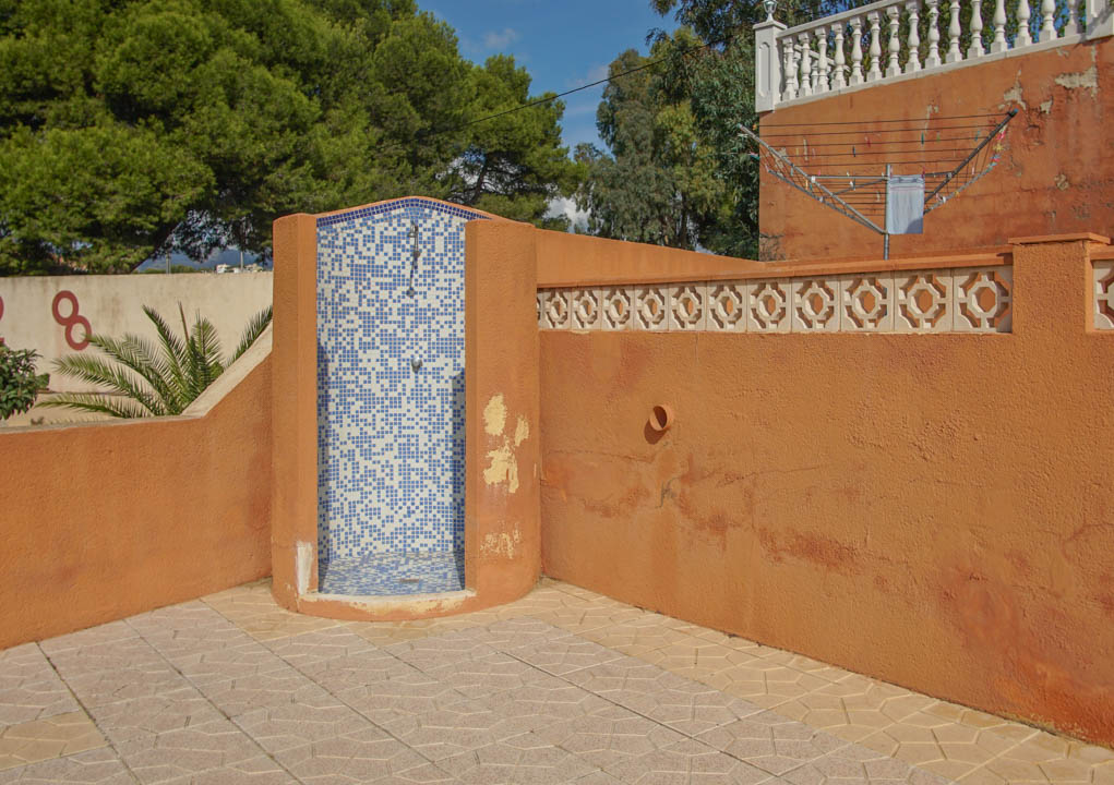 Ducha exterior revestida con mosaico azul y blanco, integrada en una zona pavimentada con muros de tono terracota y detalles decorativos de celosía.