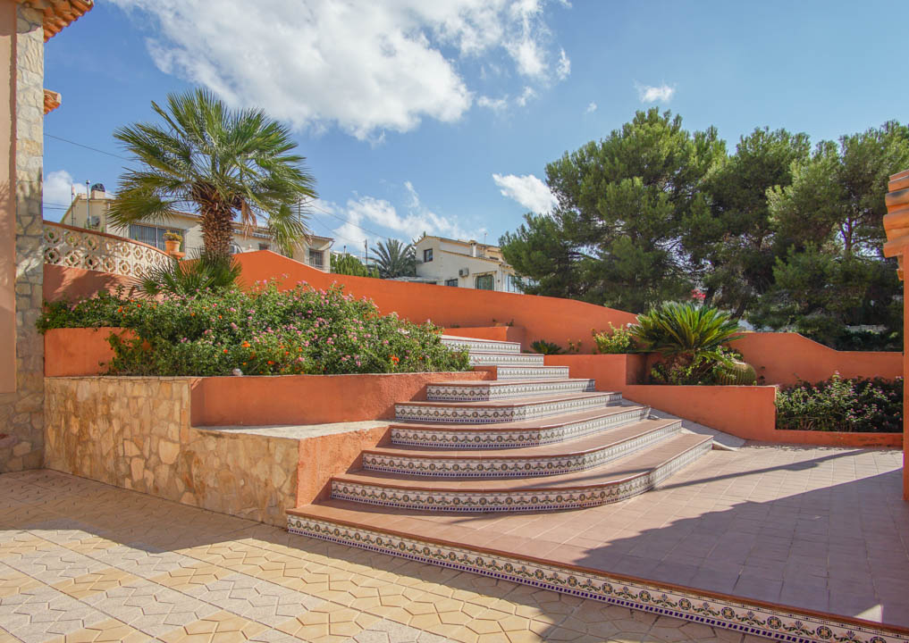 Escalera exterior con peldaños de cerámica decorada y muros en tono terracota, rodeada de jardineras con vegetación mediterránea y muros de piedra natural.