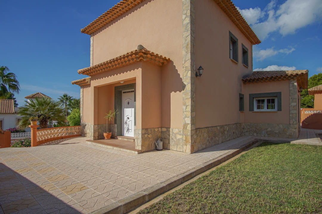 Fachada principal de la villa con entrada bajo porche, detalles en piedra natural y un amplio patio pavimentado junto a la zona de césped.