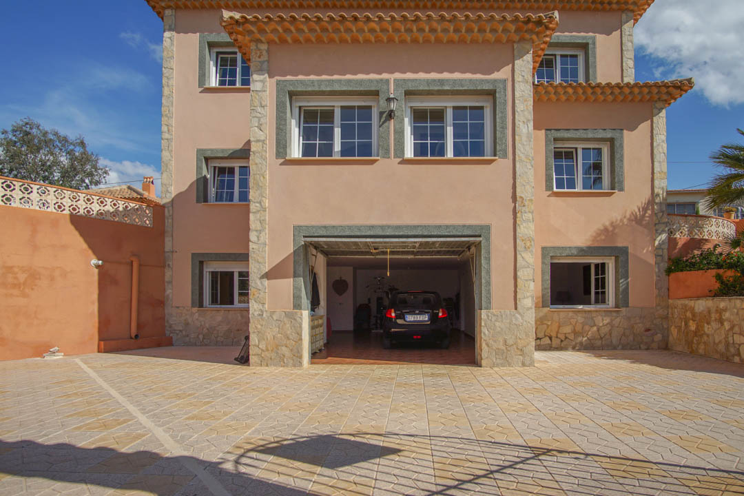 Fachada principal de tres niveles con garaje integrado, acabados en piedra natural y amplia zona de aparcamiento pavimentada con baldosas decorativas.