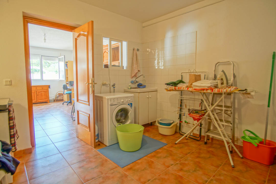 Lavadero funcional con lavadora, fregadero y zona de planchado, equipado con suelos cerámicos y acceso directo a una estancia contigua luminosa.