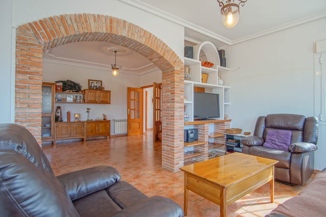 Salón con arco de ladrillo visto, suelos de gres rústico y mobiliario de madera maciza. Incluye una zona de estar con estantería de obra y acceso a otras estancias.