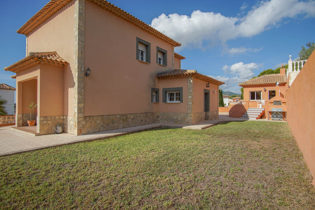 Vista exterior de la villa con fachada en tonos tierra y zócalo de piedra natural, destacando el jardín lateral y el edificio anexo con cocina exterior y horno de leña.