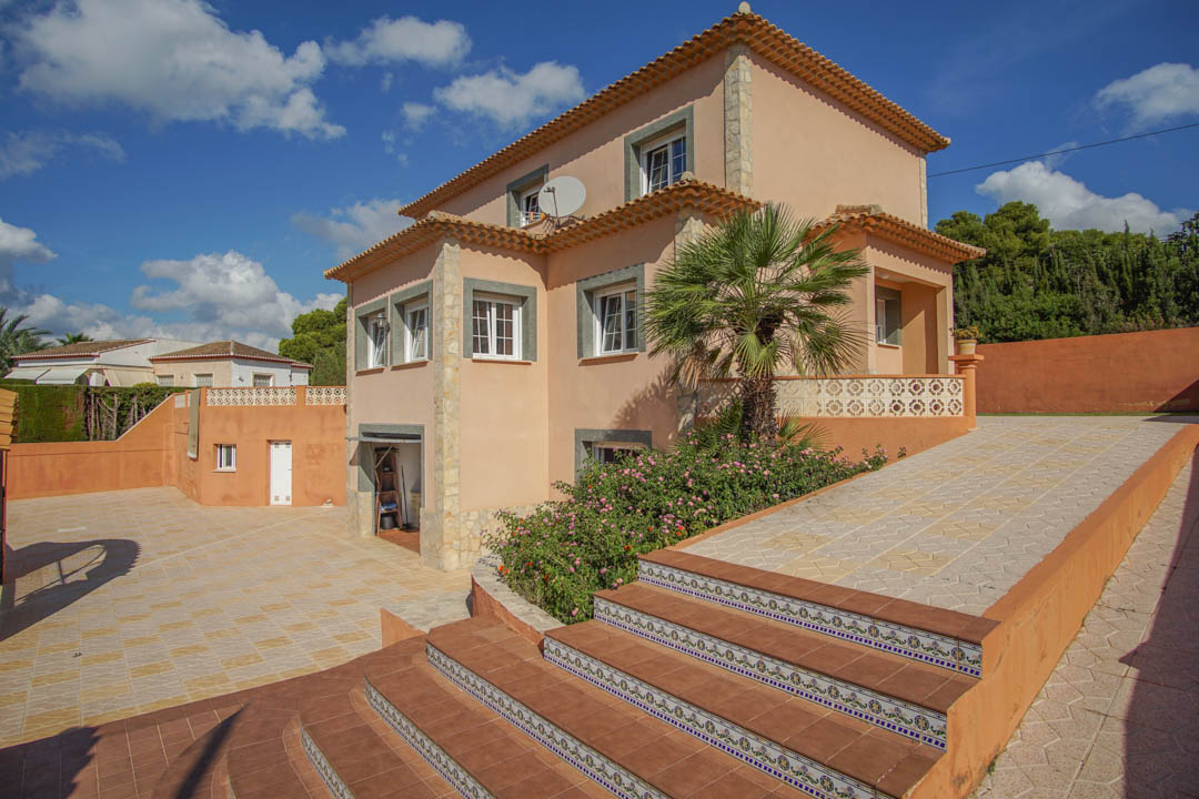 Vista exterior de la villa de tres niveles con fachada en tonos tierra, escaleras decoradas con azulejos tradicionales y un amplio patio pavimentado.
