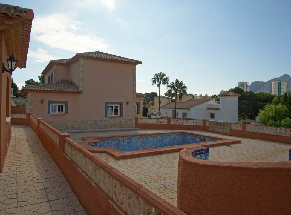 Vista exterior de la villa que muestra la amplia terraza con suelo de piedra, la piscina de mosaico azul y la arquitectura de varios niveles con tejados de teja tradicional.