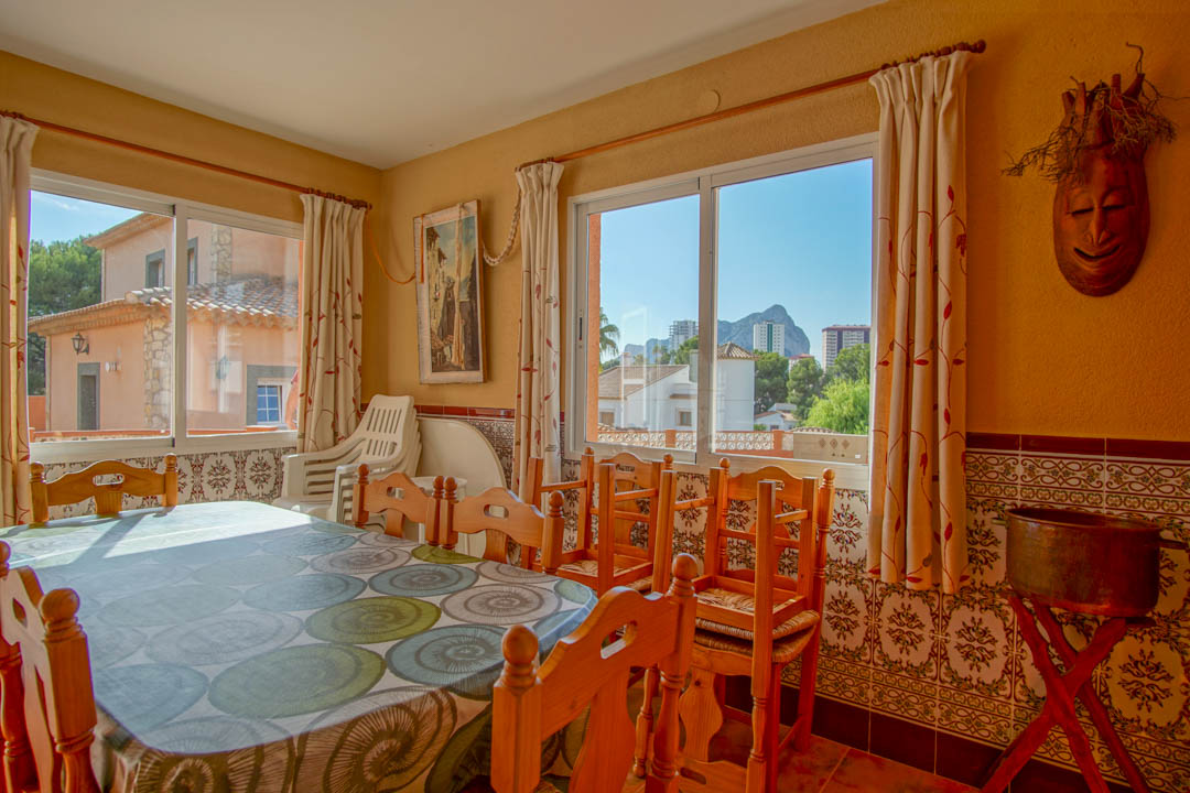 Zona de comedor luminosa con mesa de madera maciza, paredes decoradas con azulejos tradicionales y vistas al Peñón de Ifach a través de amplios ventanales.