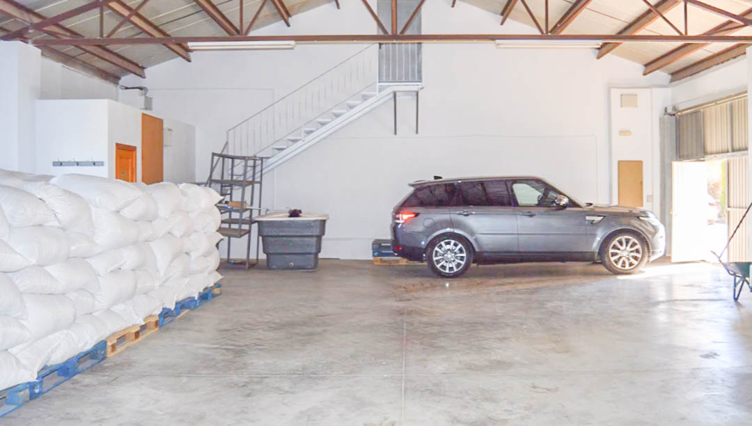 Amplio almacén industrial con suelos de hormigón, techos altos con vigas metálicas, zona de almacenamiento para sacos agrícolas y espacio para vehículos de gran tamaño.