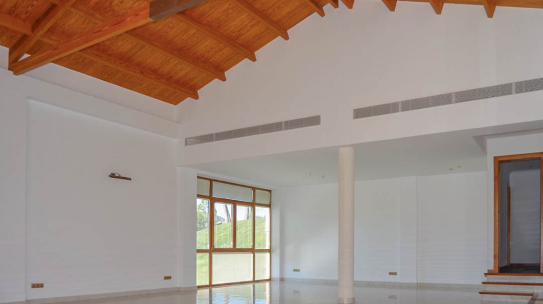 Amplio salón con techos altos de madera vista, paredes blancas y grandes ventanales que aportan luz natural. Incluye climatización integrada y suelos cerámicos pulidos.