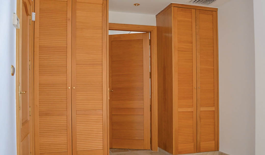 Armarios empotrados de madera maciza con puertas de lamas tipo mallorquina, integrados en un pasillo con iluminación empotrada y suelos cerámicos.