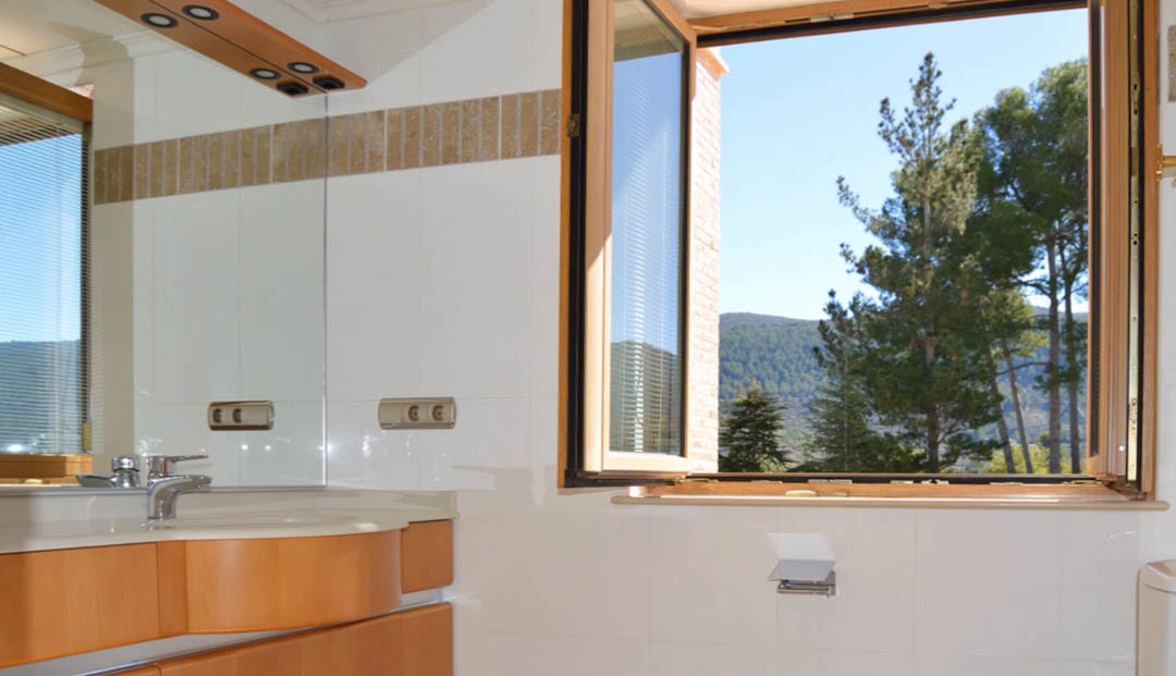 Baño luminoso con mueble de madera, encimera integrada y ventana abierta que ofrece vistas despejadas a las montañas y el bosque de pinos circundante.