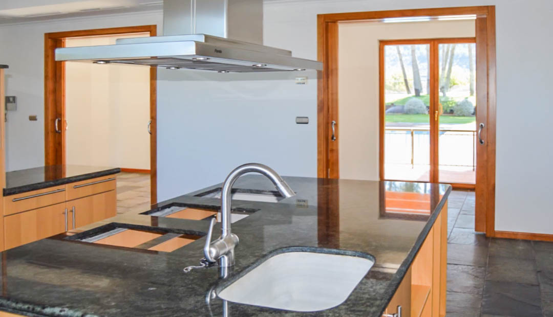 Cocina con isla de granito oscuro, campana extractora de acero inoxidable y suelos de piedra. Las puertas correderas de madera conectan con el exterior.